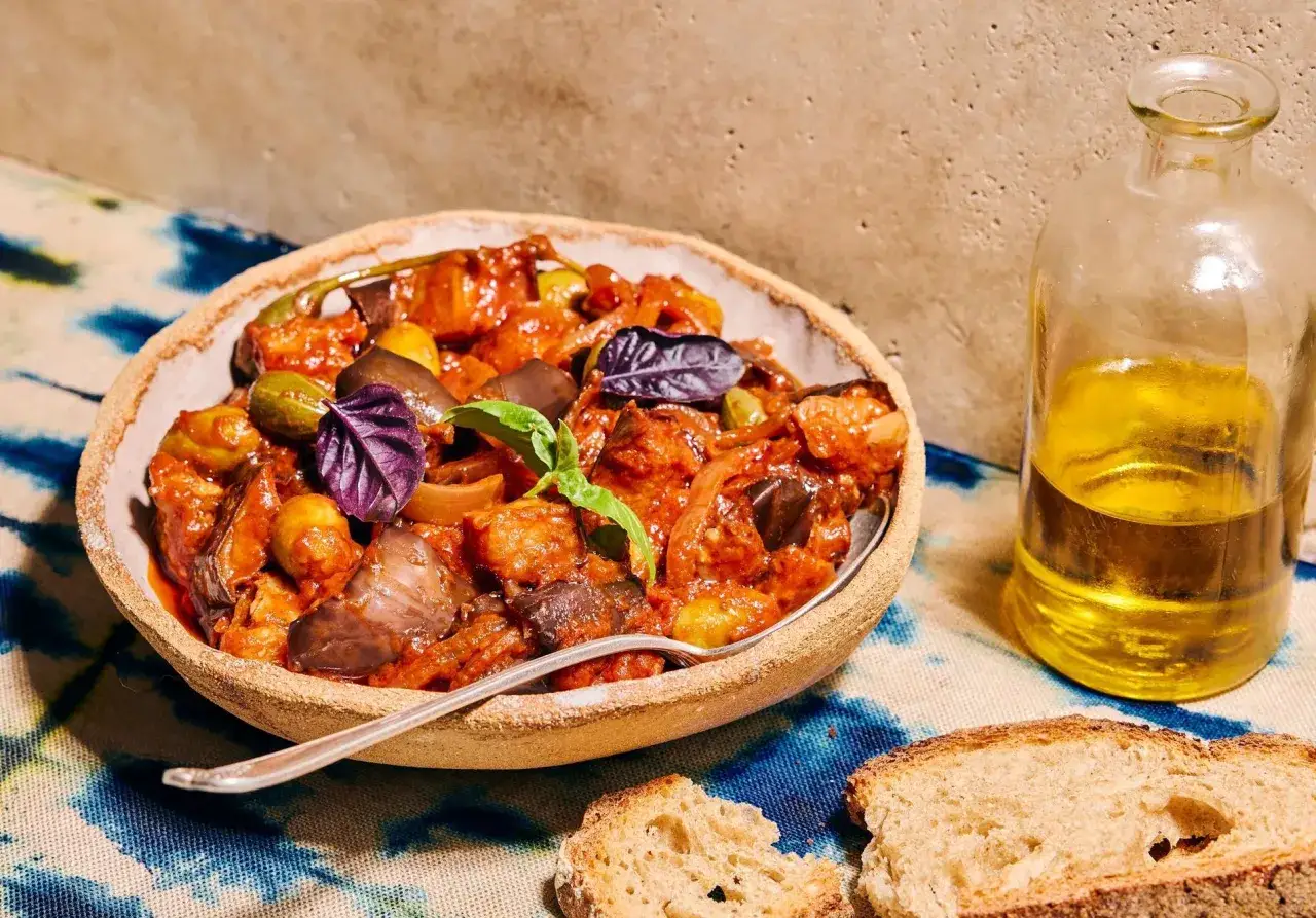 Un plat d'aubergines, olives et tomates, parfait pour un repas du soir familial rapide. Une bouteille d'huile d'olive et du pain complètent la scène.