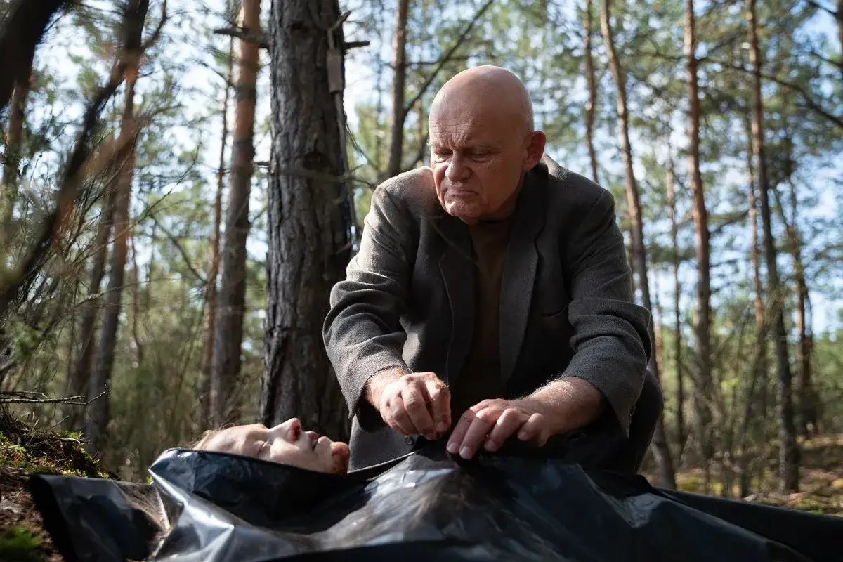W głębi lasu serial: starszy mężczyzna pochyla się nad ciałem w czarnym worku, otoczony drzewami.