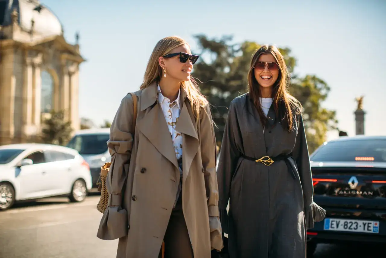Dos mujeres pasean con estilo. La de la izquierda lleva un gabán beige, uno de esos abrigos largos que llaman gabardinas. La otra, un traje gris.