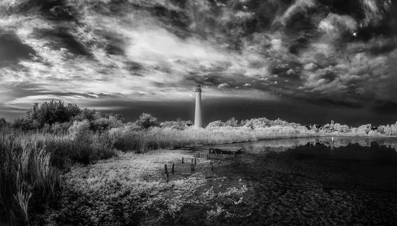 Faro blanco sobre un paisaje surrealista, con vegetación brillante y un cielo dramático. Fotografía infrarroja que revela un mundo etéreo.