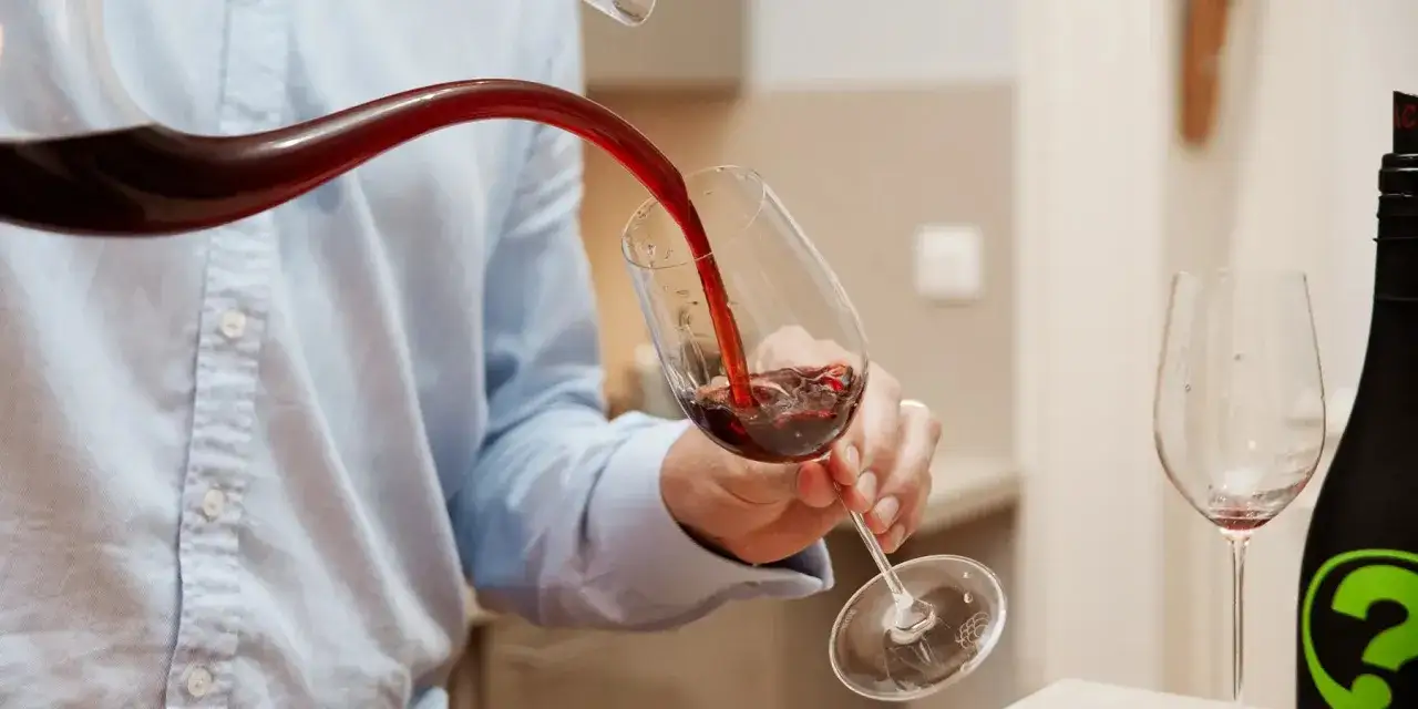 Un homme verse un vin trouble d'une carafe dans un verre. Une bouteille de vin est à côté.