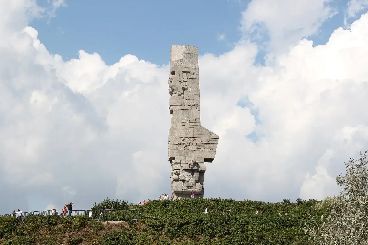 Westerplatte zwiedzanie. Monument Obrońc&oacute;w Wybrzeża g&oacute;ruje nad zielonym wzg&oacute;rzem, gdzie spacerują turyści.