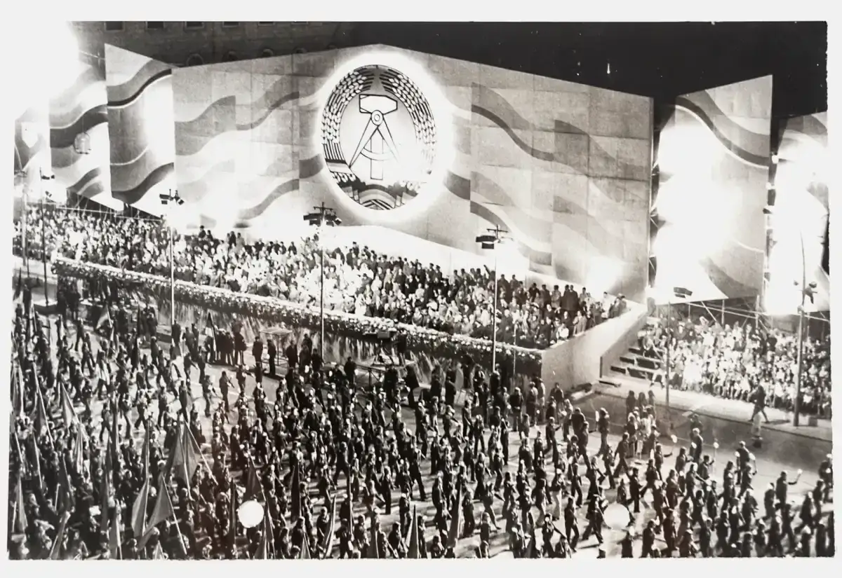 Gro&szlig;e Parade am 7. Oktober, dem Tag der Republik DDR. Tausende marschieren mit Fahnen vor einer riesigen B&uuml;hne mit dem Emblem der DDR.