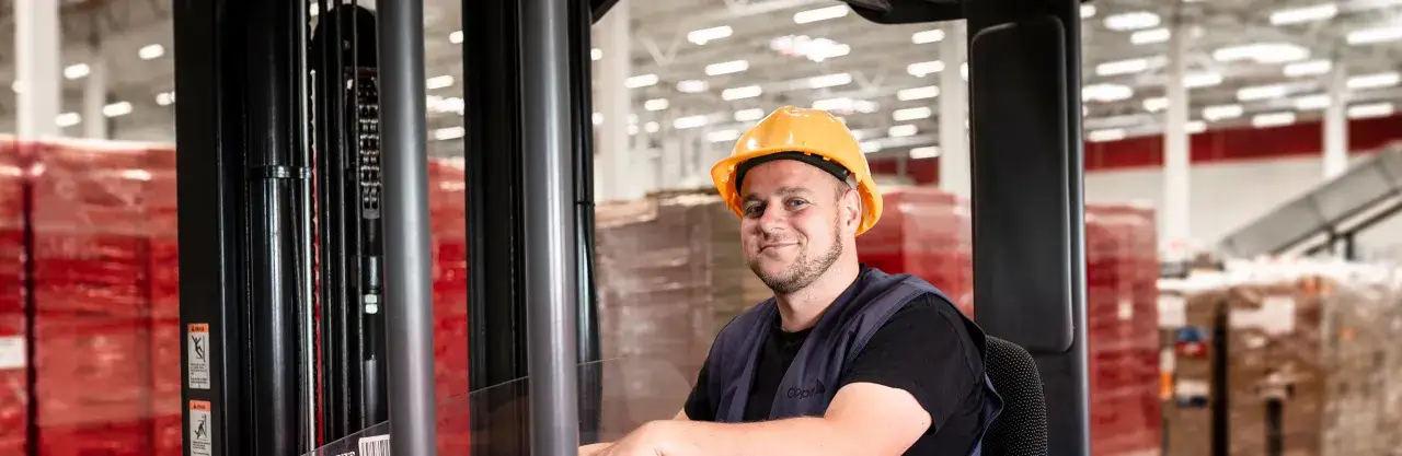 Uśmiechnięty pracownik w żółtym kasku siedzi w wózku widłowym, gotowy do pracy w magazynie. To jego praca w Limburgii.