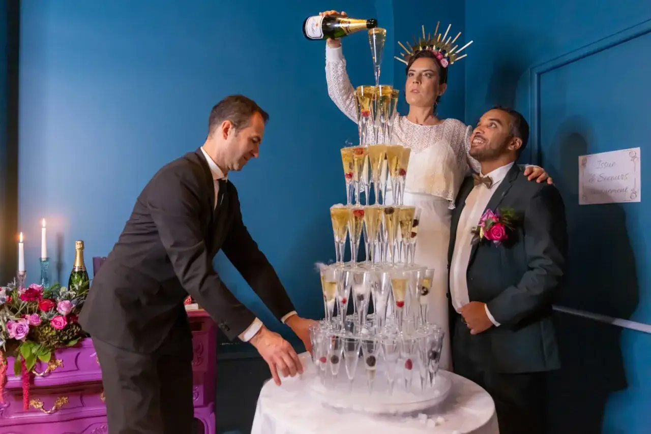 Un homme verse du champagne dans une pyramide de verres pour un vin d'honneur pour 100 personnes. La mari&eacute;e, couronn&eacute;e, et le mari&eacute; sourient.