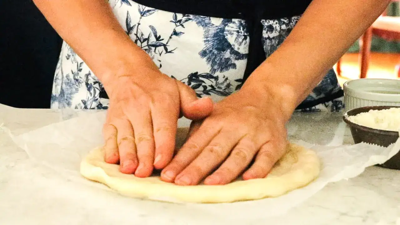 H&auml;nde kneten den pizzateig auf Backpapier. Ein Sch&auml;lchen mit geriebenem K&auml;se steht daneben.