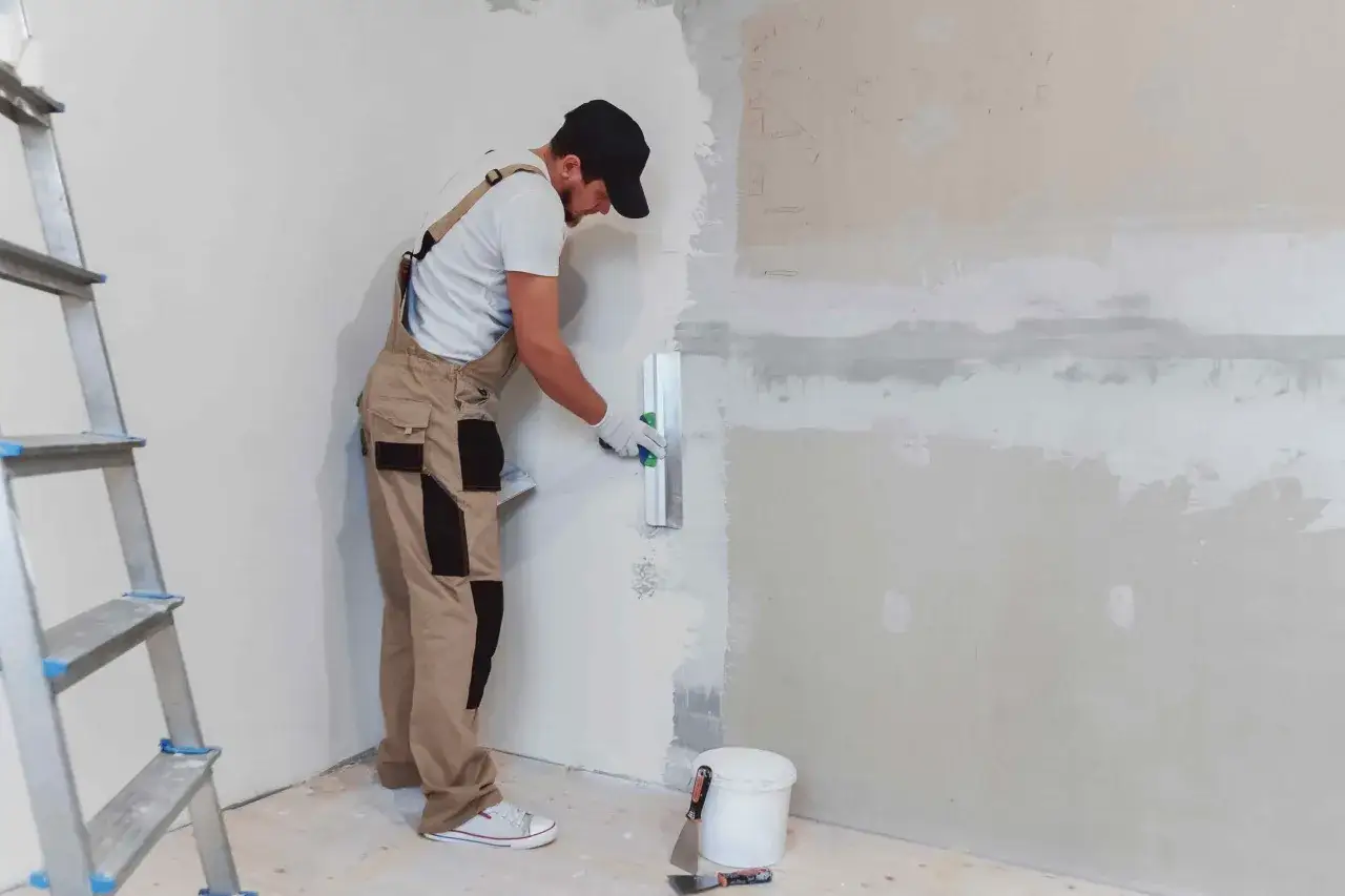 Un obrero aplica mortero sobre yeso en una pared, con una escalera y herramientas a su lado.