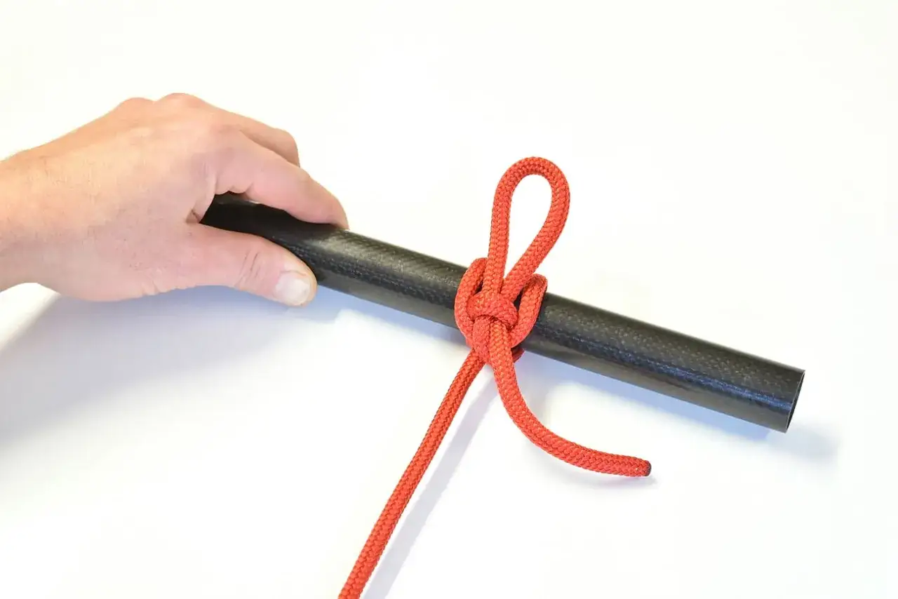 Main d'homme tenant un tube en carbone sur lequel est noué une corde rouge. Ce nœud, essentiel pour les **noeuds bateau**, est solidement fixé.