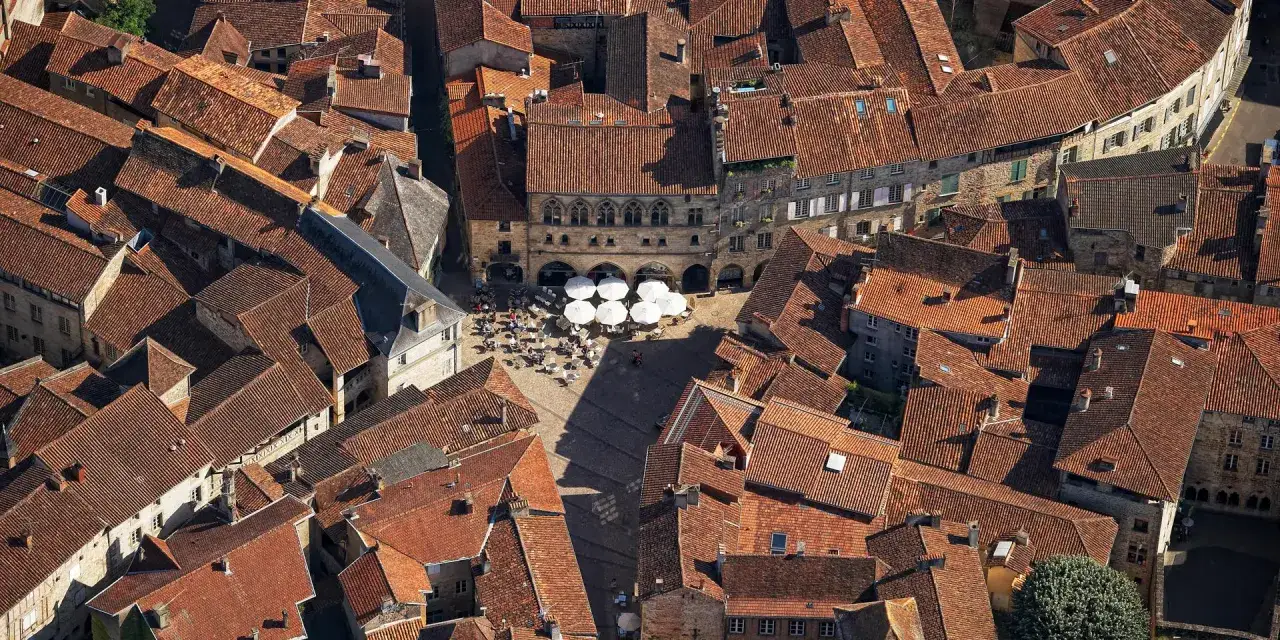 Vue aérienne des toits de tuiles rouges de Figeac, rappelant l'architecture de Cahors. Une place animée avec des parasols blancs et des terrasses.