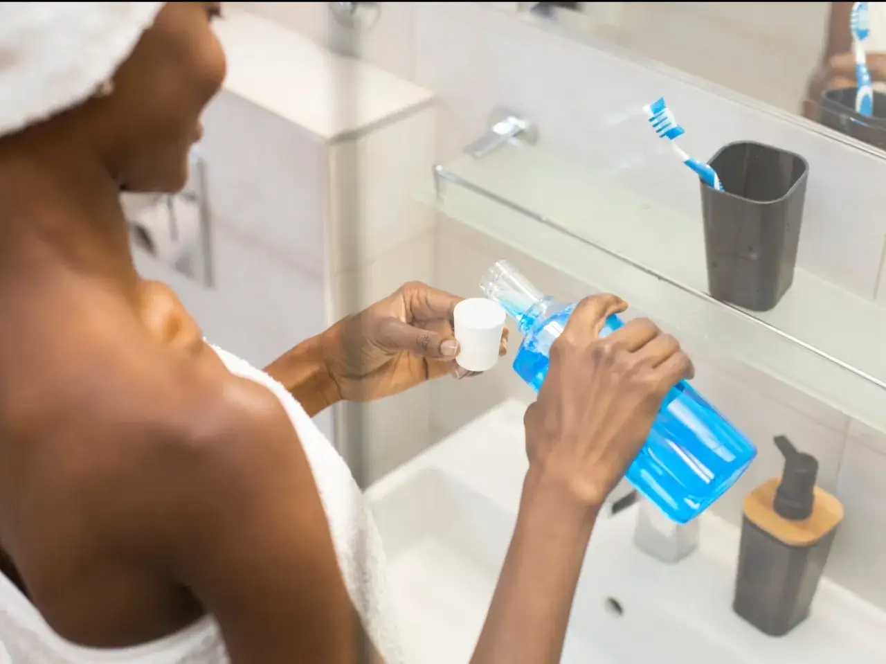 Kobieta w ręczniku nalewa niebieską płukankę do zęb&oacute;w do białego kubeczka.