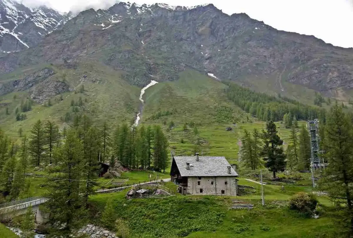 Un casolare in pietra nel **Val di Rhemes**, circondato da verdi pendii e imponenti montagne innevate.