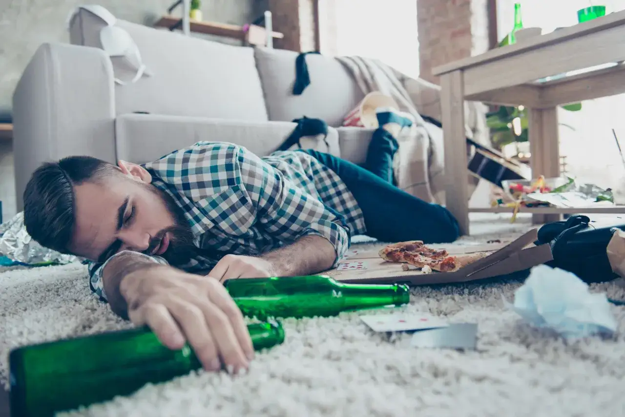 Mężczyzna śpi na dywanie, otoczony pustymi butelkami po piwie, resztkami pizzy i kartami. To obrazek pokazujący skutki nadużywania alkoholu, być może padaczka alkoholowa po jakim czasie picia.