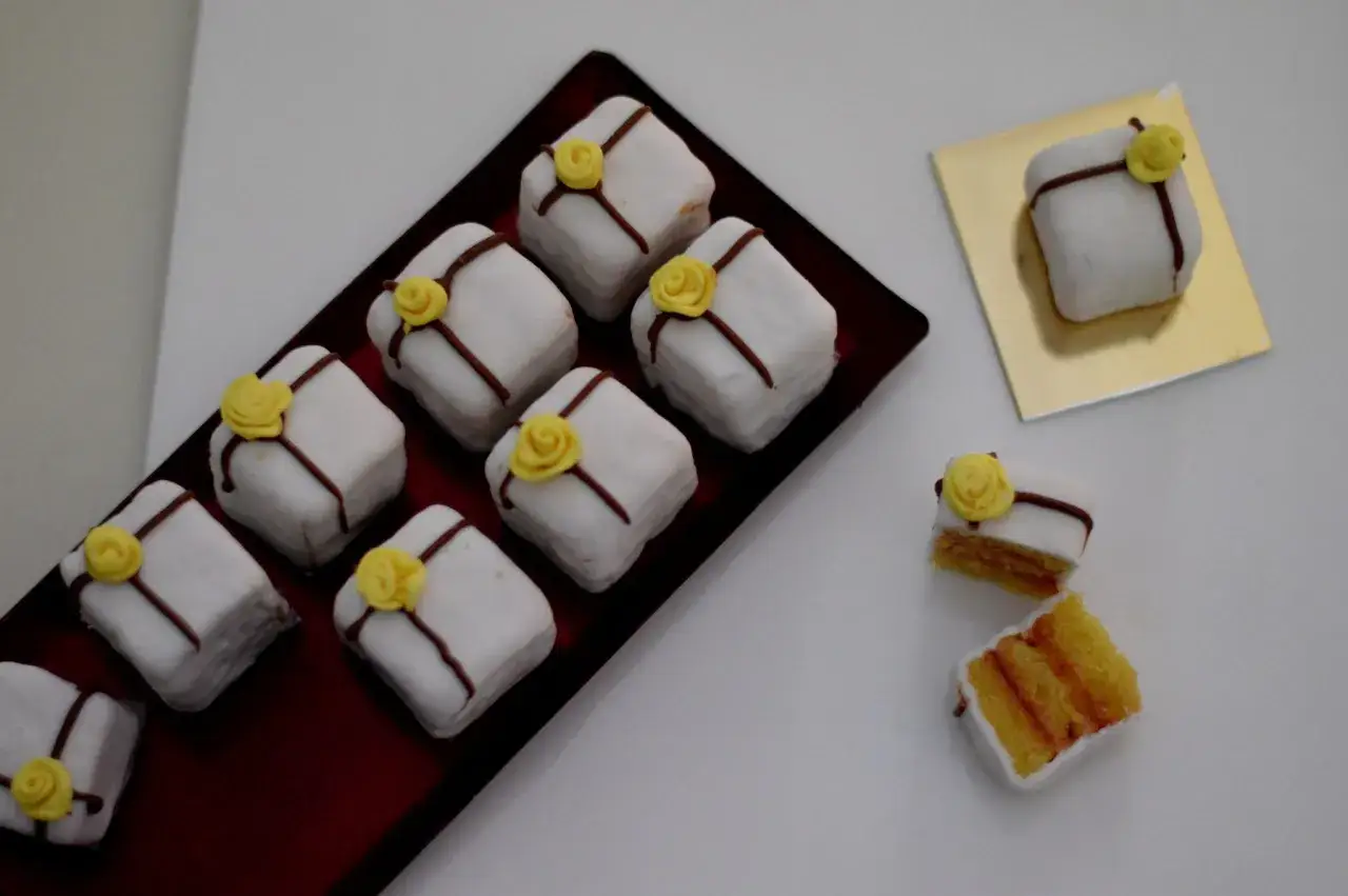Petit four que es: peque&ntilde;os pasteles cuadrados cubiertos de glaseado blanco, decorados con una rosa amarilla y l&iacute;neas de chocolate.
