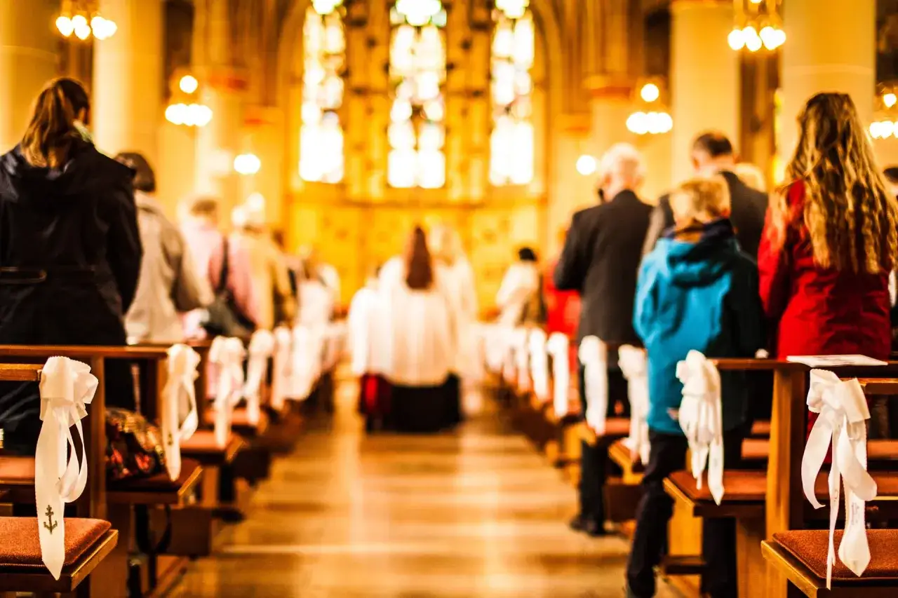 Menschen sitzen in einer Kirche, die Bänke sind mit weißen Schleifen geschmückt. Ein Kind im blauen Hoodie steht im Gang. Der **apostolische Glaubensbekenntnis** wird vielleicht gerade rezitiert.