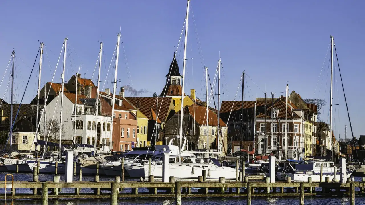 Segelboote im **Flensburg Hafen** vor bunten H&auml;usern und einem Kirchturm unter blauem Himmel.