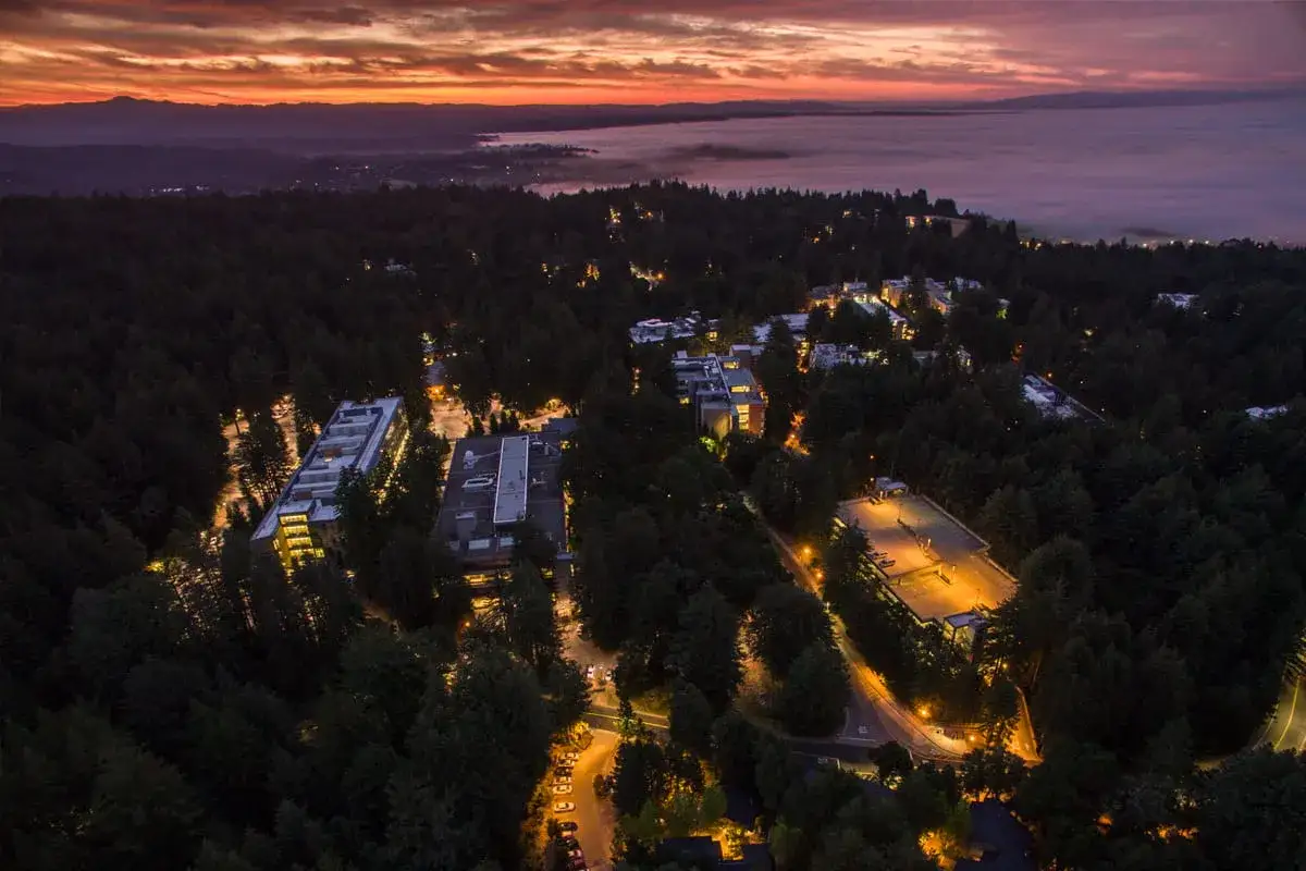 Vue a&eacute;rienne de nuit sur le campus de UC Santa Cruz, illumin&eacute; par des lumi&egrave;res chaudes, avec un ciel orang&eacute; et une mer de brouillard au loin.
