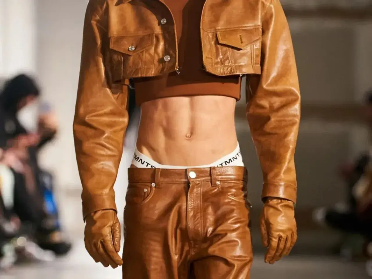 A runway model showcases a bold look with a cropped leather jacket, revealing toned abs and branded underwear. This daring ensemble, featuring leather pants and gloves, is a statement for gay men in sportswear.