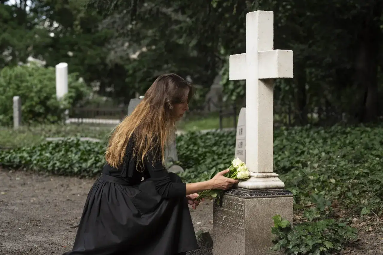 Kobieta w czarnej sukni klęczy przy grobie, kładąc białe róże. Modlitwa 