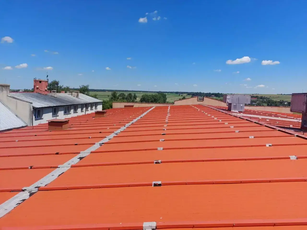 Pomarańczowy dach z minimalnym spadkiem dachu płaskiego, widok na pola i niebo.