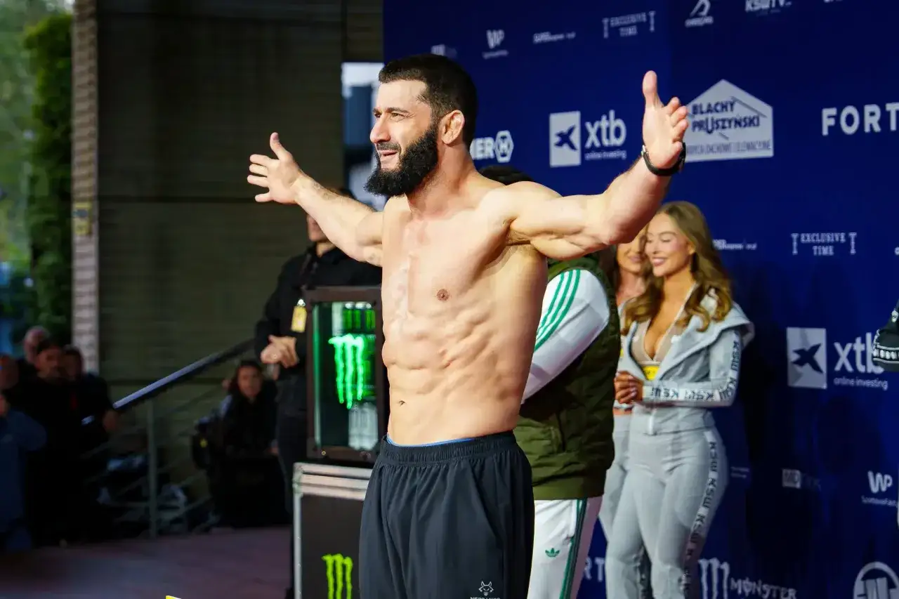 KSW relacja: muskularny zawodnik z brodą pozuje z rozłożonymi rękami na tle niebieskiej ścianki z logotypami sponsorów.