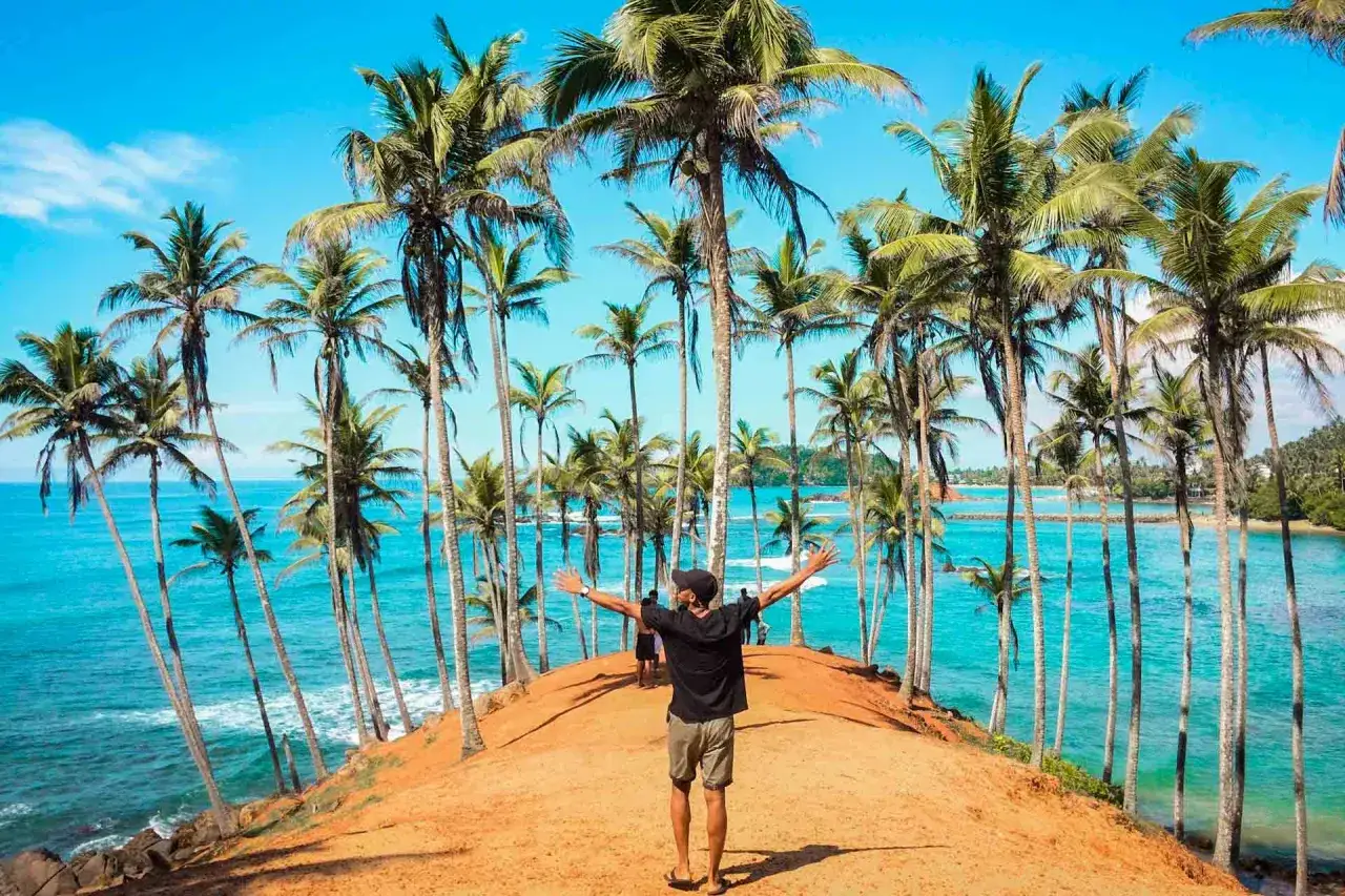 En man med utsträckta armar njuter av den fantastiska utsikten över palmer och turkost hav på Sri Lanka.