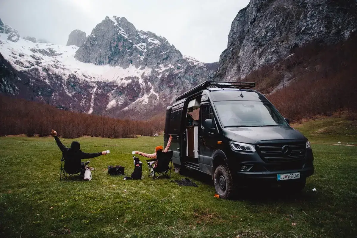 Zwei Personen und zwei Hunde genießen eine Pause auf ihrer Montenegro-Route, umgeben von majestätischen Bergen und schneebedeckten Gipfeln.