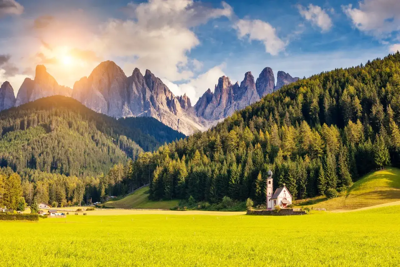 En solig sommardag i Dolomiterna. En liten kyrka st&aring;r p&aring; en gr&ouml;n &auml;ng framf&ouml;r en t&auml;t granskog och dramatiska bergstoppar.