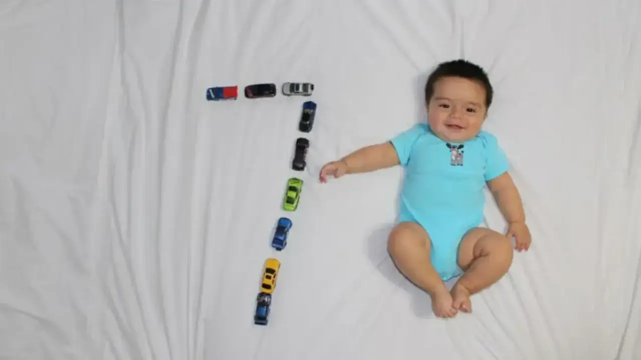 Un feliz bebe de 7 meses sonr&iacute;e junto a una fila de coches de juguete que forman el n&uacute;mero siete.