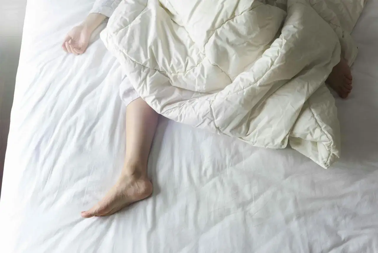 En person sover i en vit s&auml;ng, t&auml;ckt av ett vitt t&auml;cke. En arm och ett ben syns. Kanske upplever personen svettningar gravid.