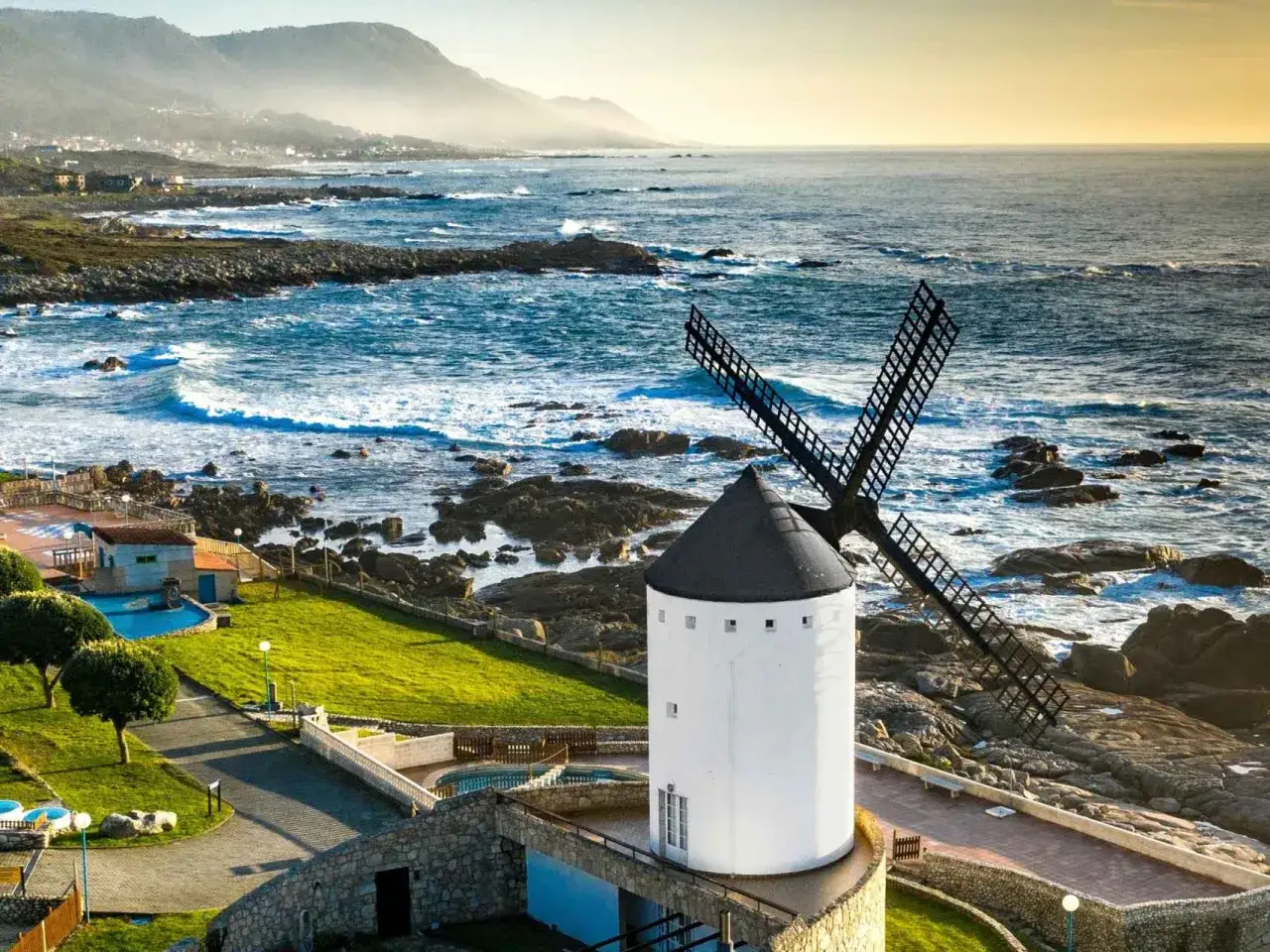 Un mui&ntilde;o do vento blanco con aspas negras se alza junto a un mar azul y rocoso. El sol ilumina la costa y las monta&ntilde;as al fondo.