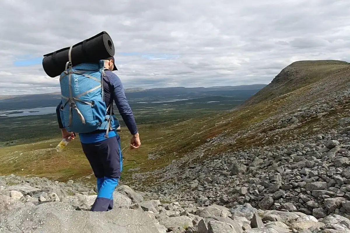 Vandrare med stor ryggsäck och liggunderlag på väg nerför en stenig fjällsida. Utsikt över sjöar och skog. Perfekt för mat och vandring.