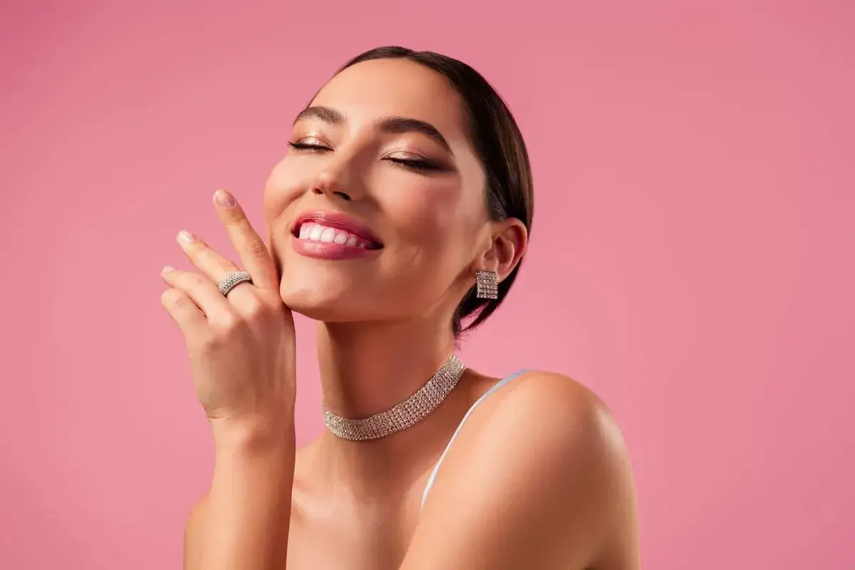 Mujer sonríe con ojos cerrados, luciendo un maquillaje romántico, joyas brillantes y un fondo rosa.