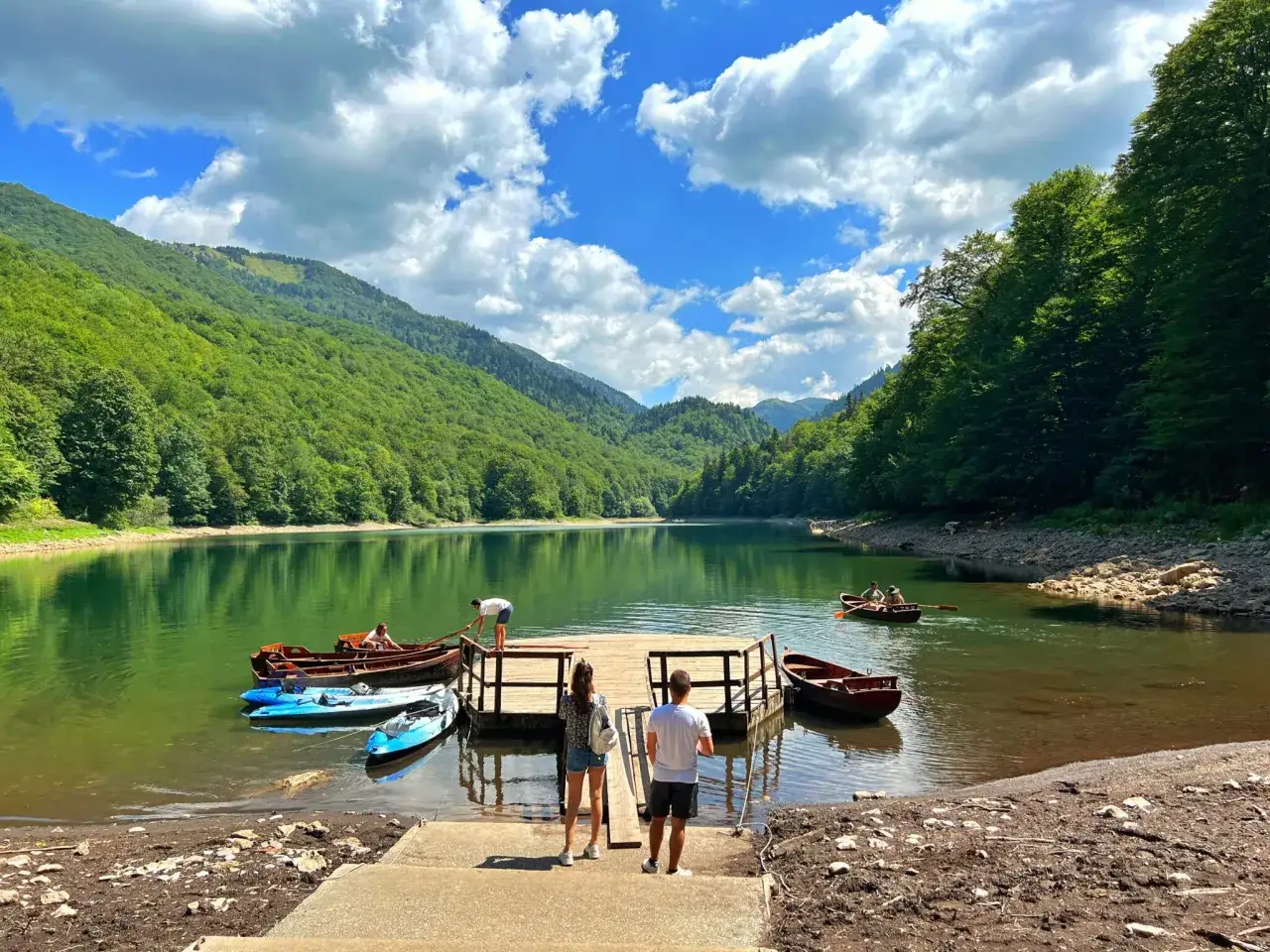 Malownicze jezioro otoczone zielonymi wzgórzami. Drewniane pomosty, łodzie i kajaki czekają na wodną przygodę. Idealne miejsce na camping w Czarnogórze.