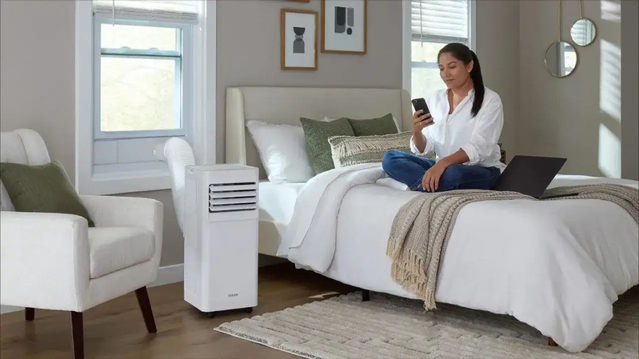 Une femme utilise son téléphone sur un lit, à côté d'un climatiseur mobile, la solution idéale pour un confort optimal.