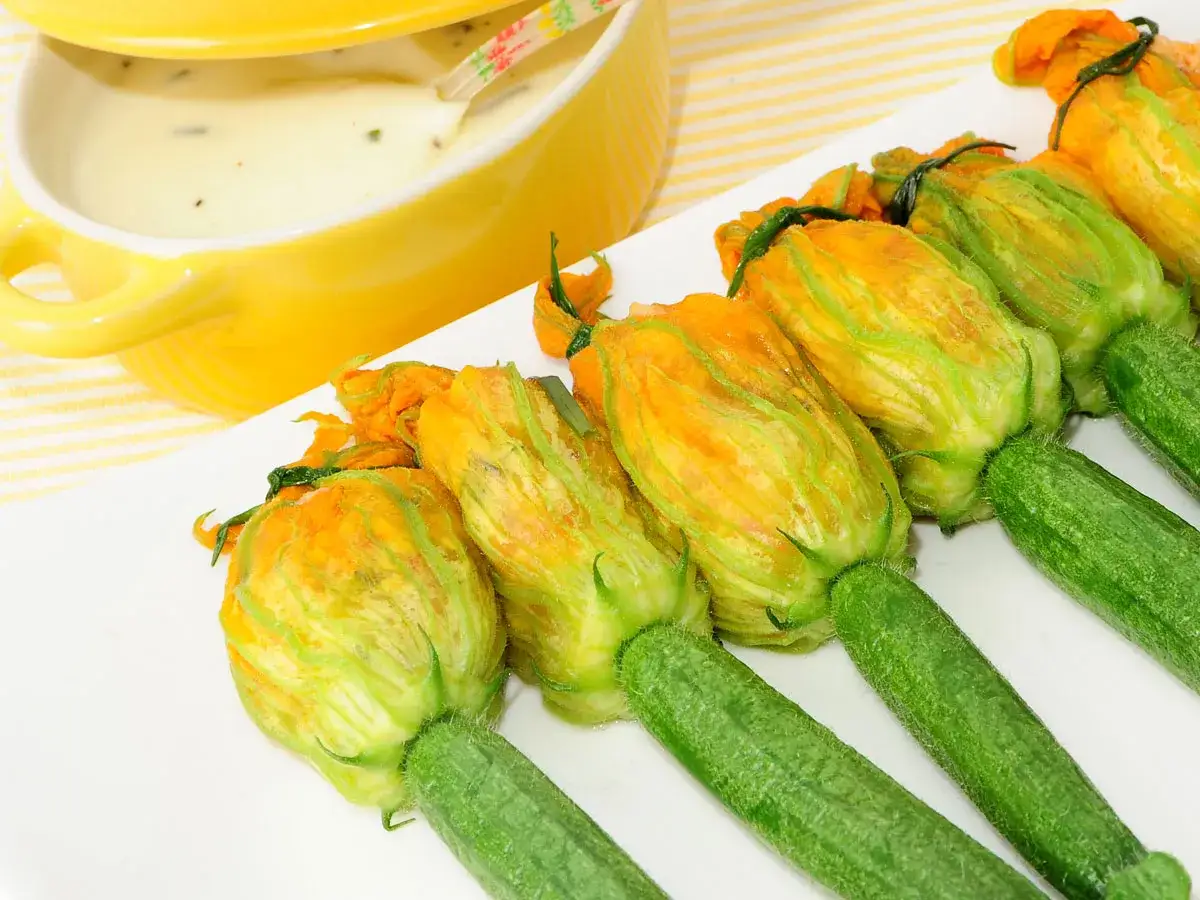 Deliciosos rellenos de flor de calabac&iacute;n listos para fre&iacute;r, acompa&ntilde;ados de una cremosa salsa en un cuenco amarillo.