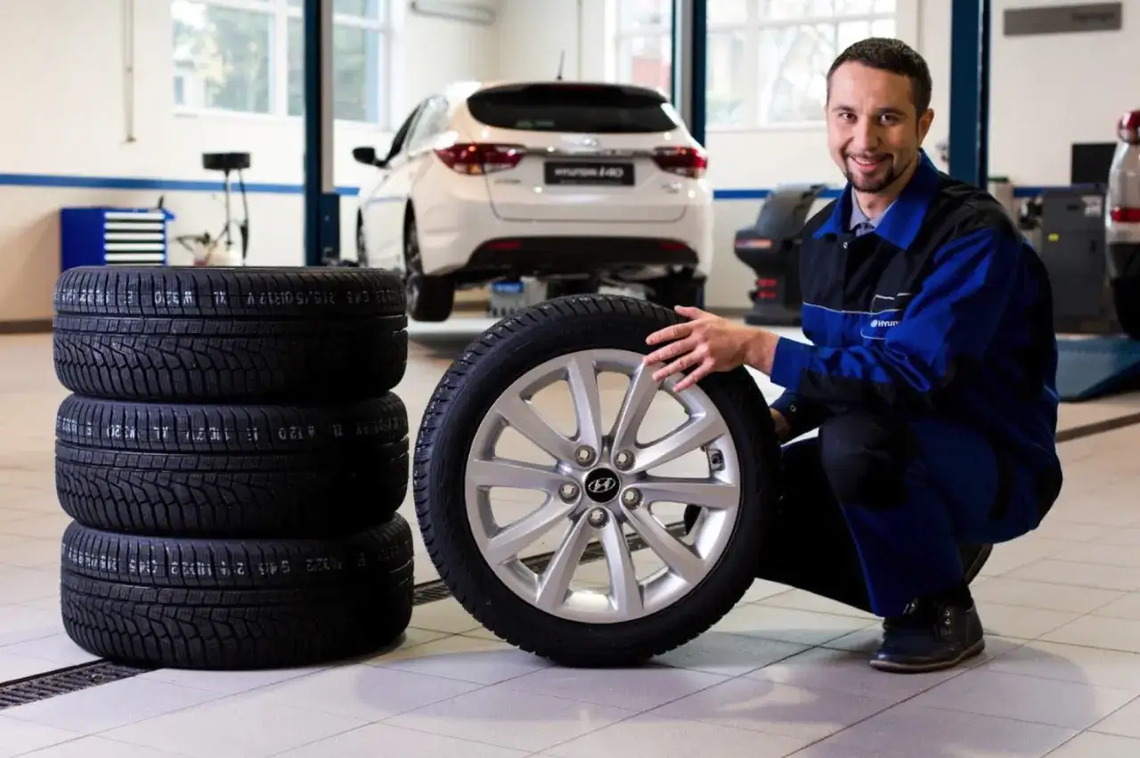 Uśmiechnięty mechanik prezentuje komplet opon zimowych gotowych do zmiany. W tle biały Hyundai i40.