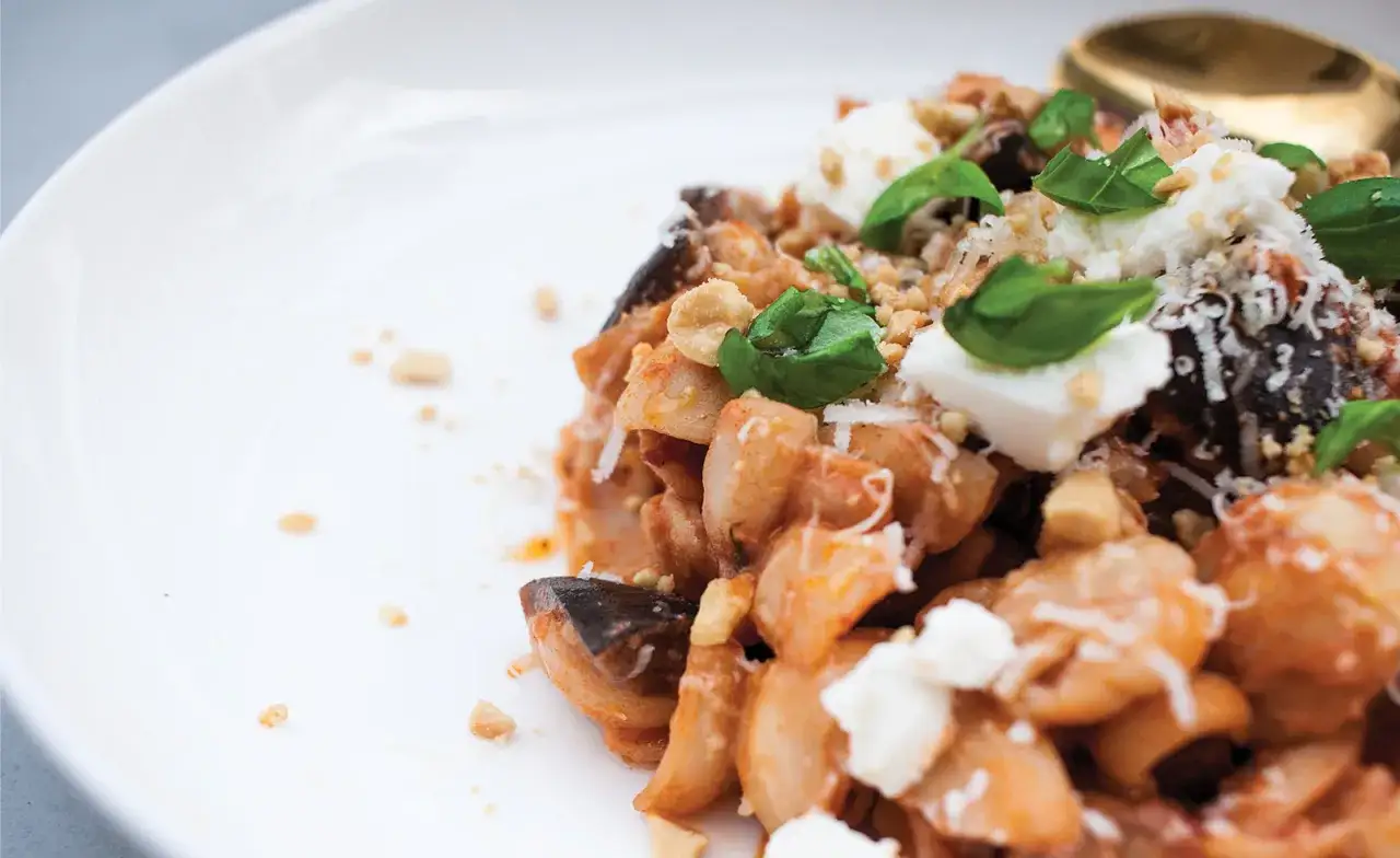 Pyszne danie pasta alla norma z bakłażanem, pomidorami i fetą, ozdobione bazylią i orzeszkami.