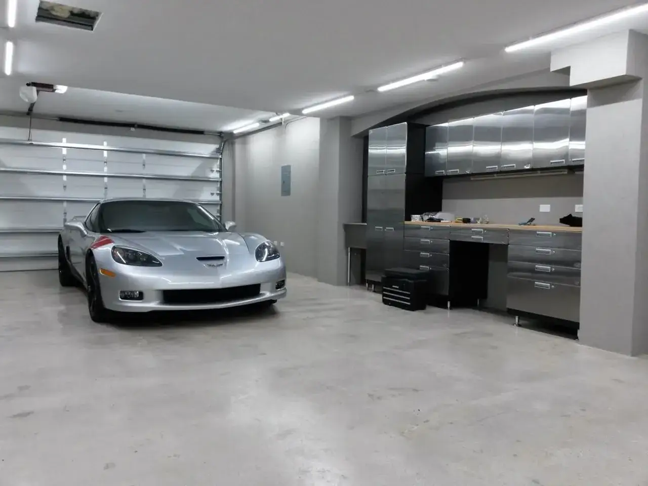 Srebrny Chevrolet Corvette stoi na gładkim, polerowanym betonie w garażu z metalowymi szafkami i jasnym oświetleniem.