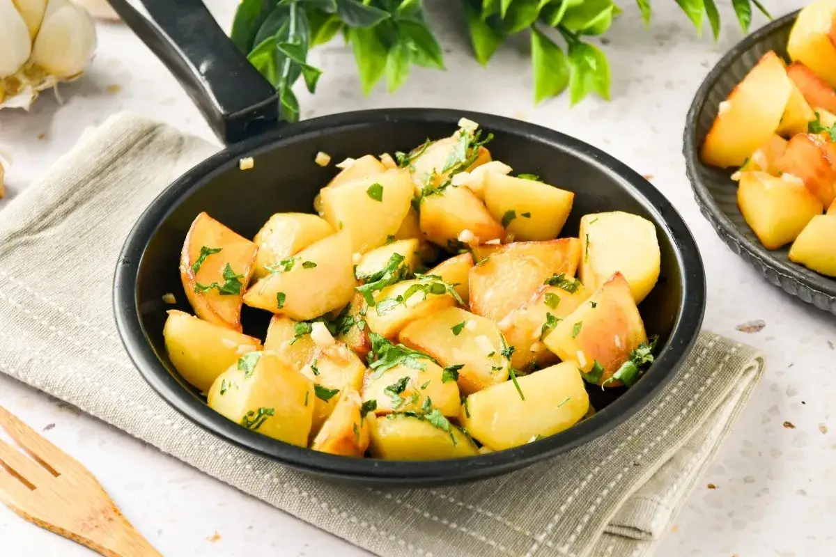 Patatas al ajillo recién hechas, doradas y salteadas con ajo y perejil fresco, servidas en una sartén negra.