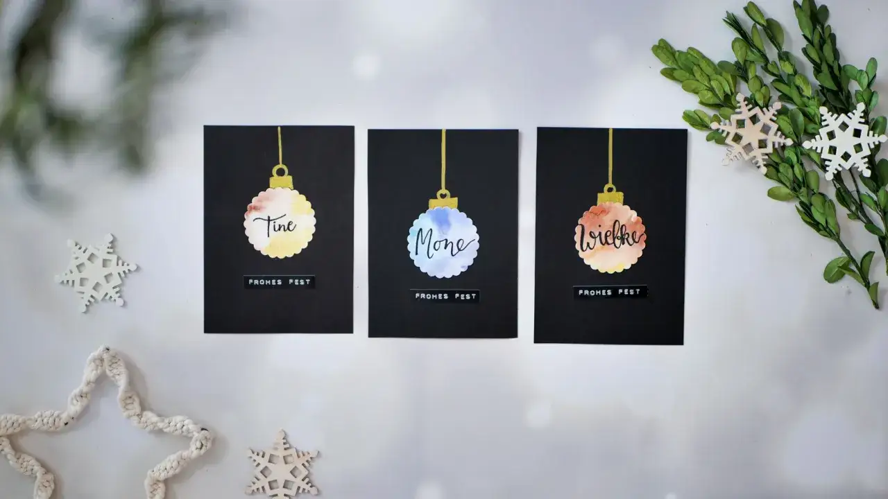 Moderne weihnachtskarten basteln: drei schwarze Karten mit Aquarell-Christbaumkugeln und Namen. Dekoriert mit Schneeflocken und Gr&uuml;n.