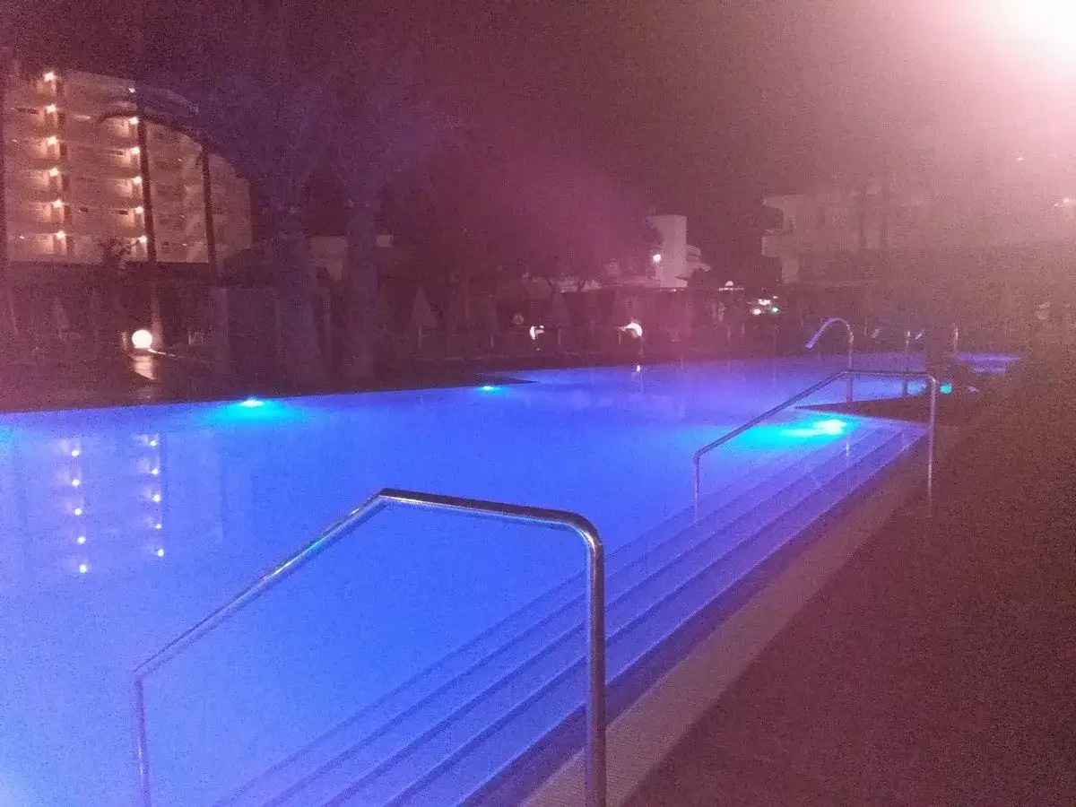 Nocny basen z niebieskim podświetleniem, schodkami i poręczami. W tle widać budynek hotelowy, prawdopodobnie Hipotels Gran Coma.