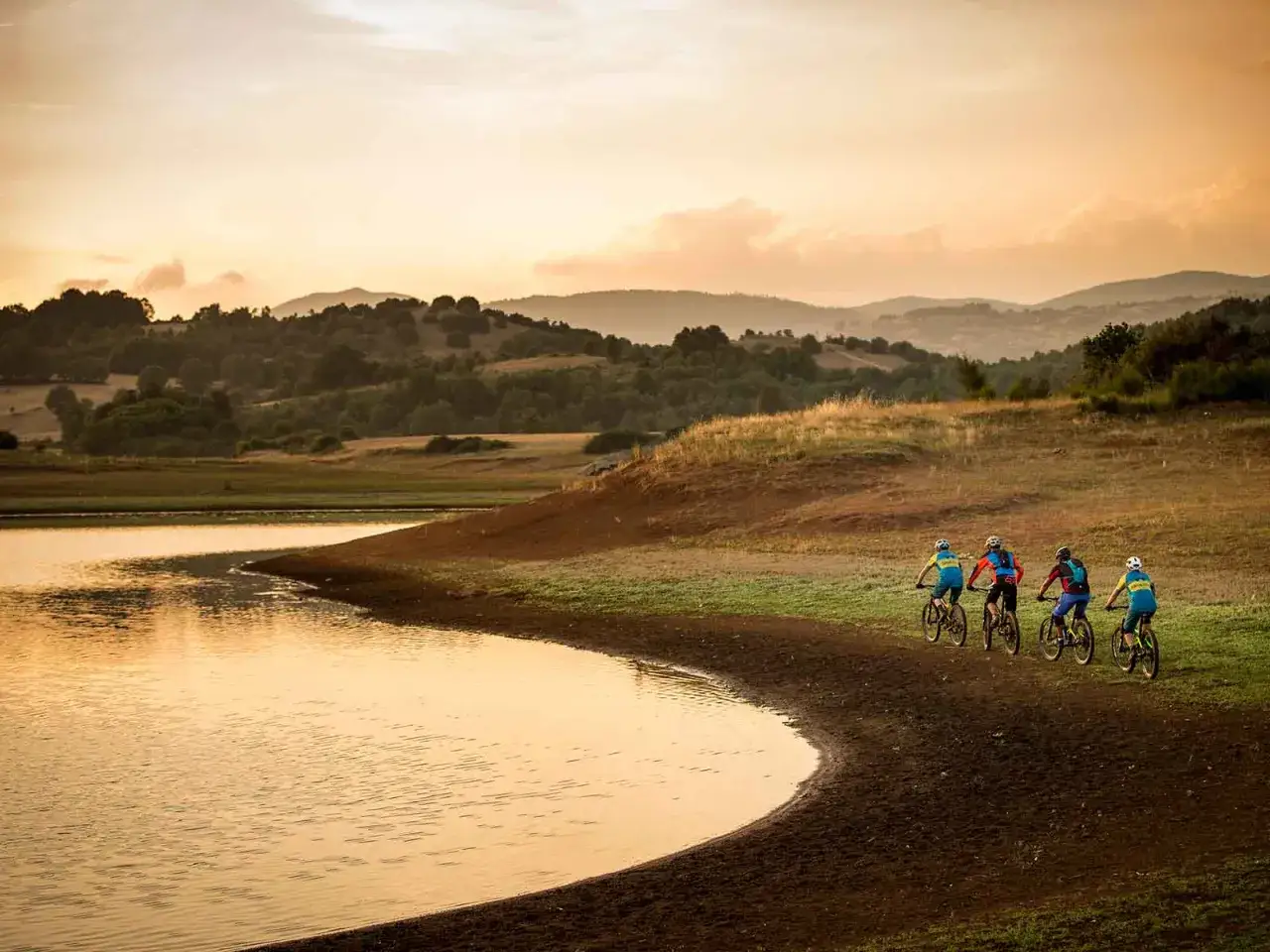 Quattro ciclisti pedalano lungo la riva del lago di Canterno al tramonto, con dolci colline sullo sfondo.