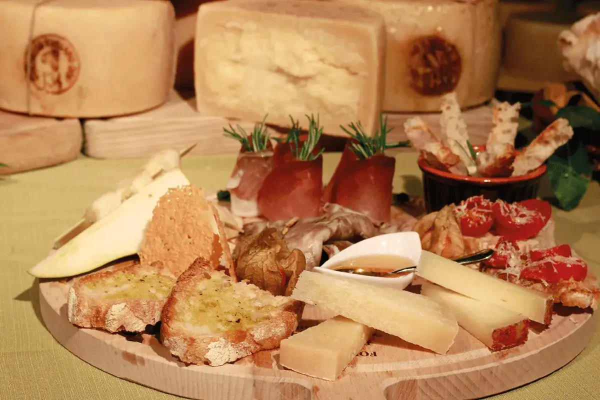 Assaggio di formaggi grassi quali sono: stagionati, freschi, con salumi, pane e pomodorini su tagliere di legno.