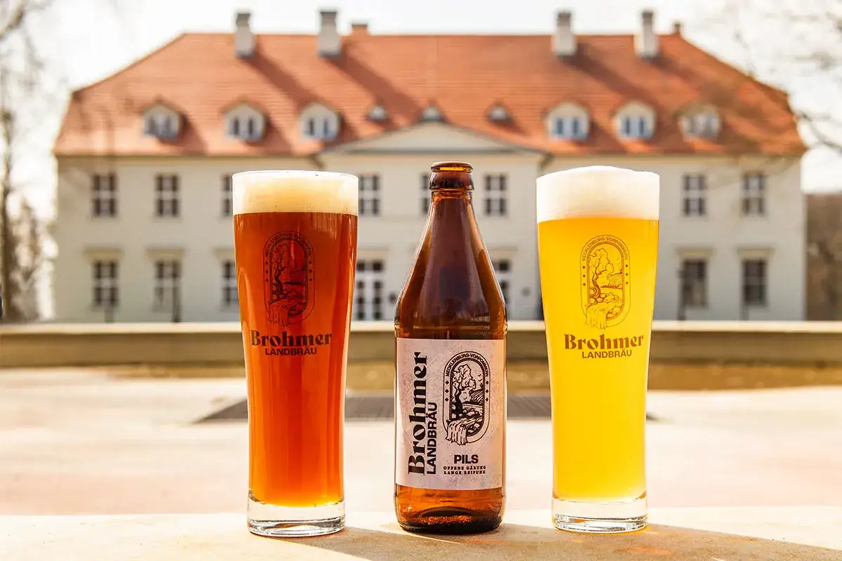 Zwei Gläser Brohmer Landbräu, eines bernsteinfarben, das andere hellgelb, stehen neben einer Flasche. Das unfiltriertes Bier ist bereit zum Genuss vor einem Schloss.