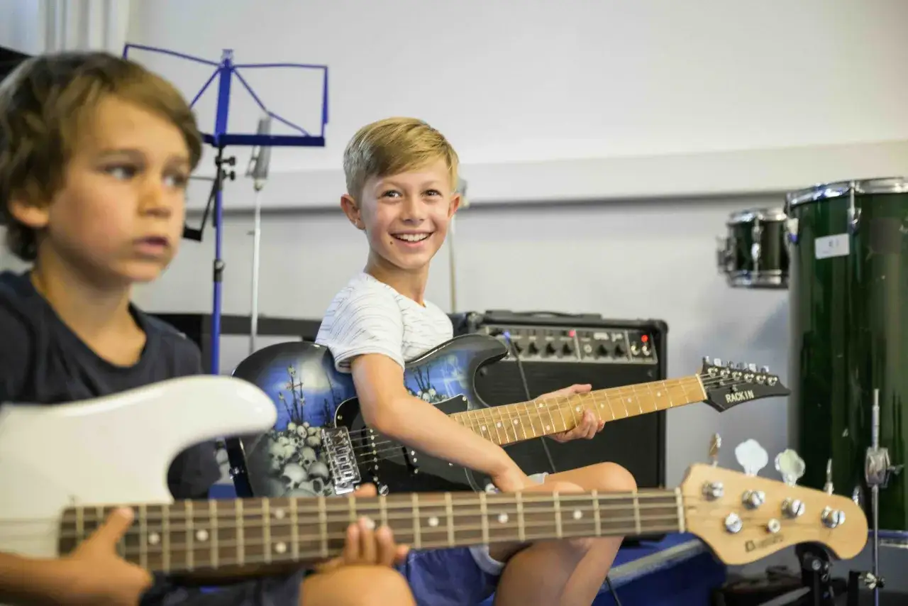 Zwei Jungen &uuml;ben in der Musikschule Gitarre. Einer l&auml;chelt fr&ouml;hlich in die Kamera, w&auml;hrend der andere konzentriert auf seine Gitarre blickt.