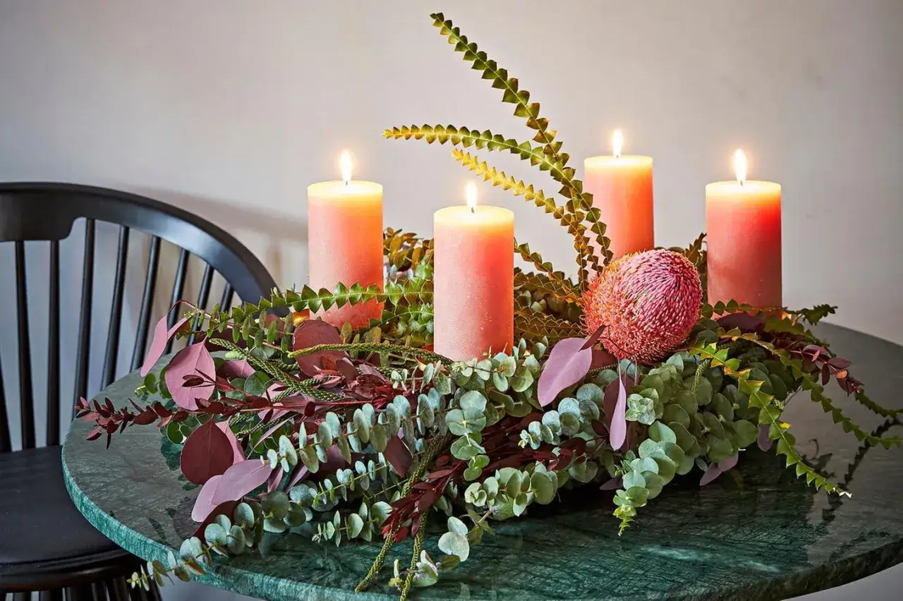 Ausgefallener Adventskranz Trend: Vier pinke Kerzen umgeben von Eukalyptus, roten Blättern und einer Banksia-Blüte auf einem grünen Marmortisch.
