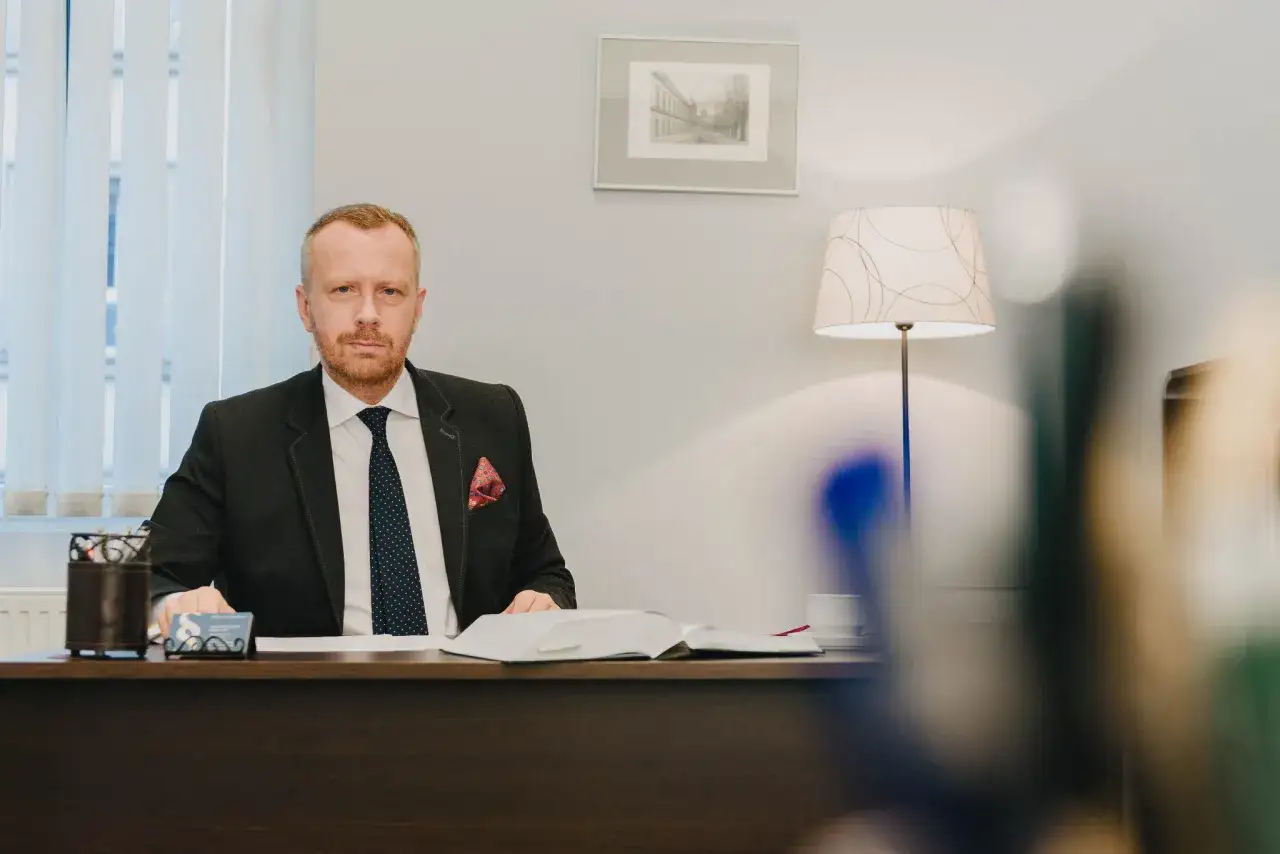 Mężczyzna w garniturze siedzi przy biurku, przed nim otwarta księga i wizytówka.