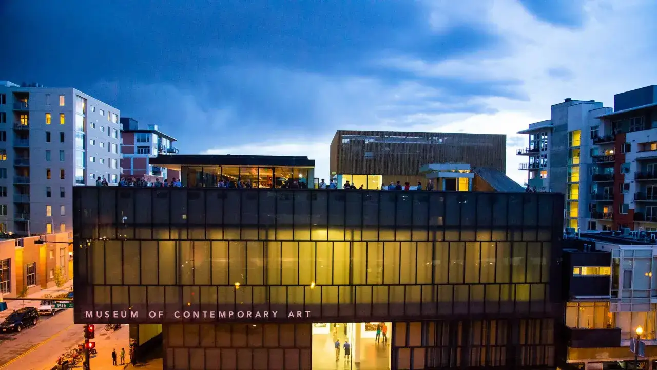 Museum of Contemporary Art Denver photos: A modern building with glass facade, people on the rooftop, and surrounding city buildings at dusk.