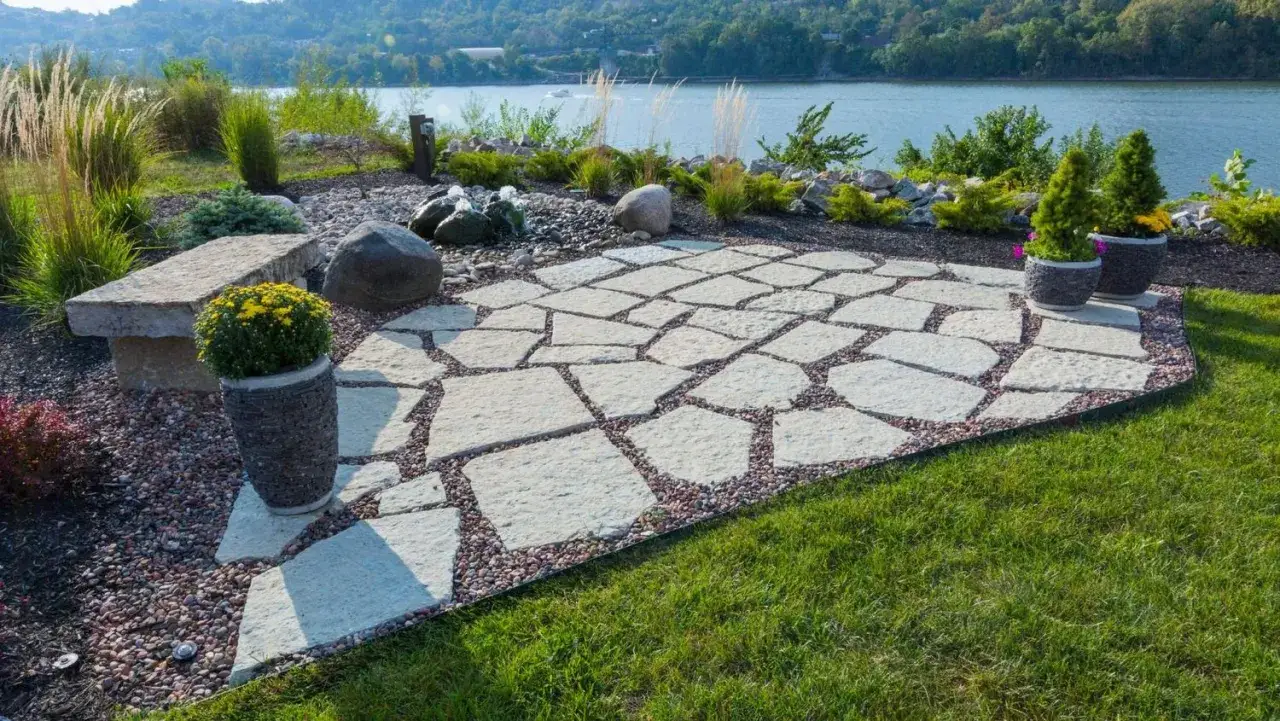 Une belle terrasse en pav&eacute; irr&eacute;gulier, bord&eacute;e de gravier, avec un banc en pierre et des plantes, surplombant une rivi&egrave;re paisible.