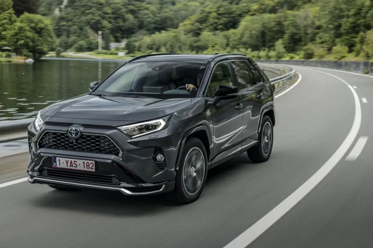 Ciemnoszary Toyota RAV4 2025 pokonuje zakręt na malowniczej drodze nad jeziorem.